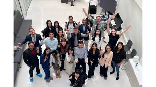 Looking down onto a group of Postgraduate students standing in a foyer raising their arms and hands posing and smiling towards the camera